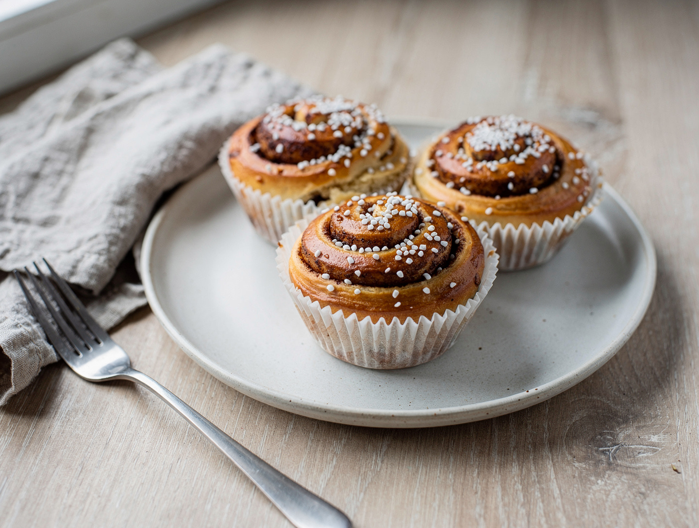 Tre kanelbullar med pärlsocker i pappersformar på en off-white tallrik